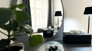 Ein Hotelzimmer mit rundem Spiegel, einer Topfpflanze, einem Nachttisch mit Pflegeutensilien, einem schwarzen Hocker und einer Lampe.