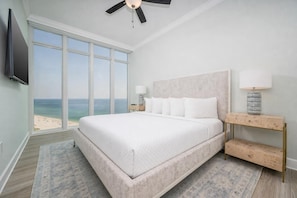 A bedroom with a large bed, a ceiling fan, a TV, a nightstand with a lamp, and a view of the beach.