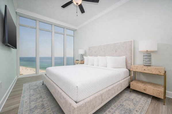 A bedroom with a large bed, a ceiling fan, a TV, a nightstand with a lamp, and a view of the beach.