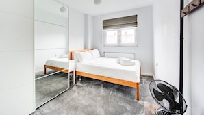 A modern bedroom with a large mirror, a bed with white linens, a window with blinds, and a standing fan. - GuestReady - Trendy 2Bed flat in East London (London)