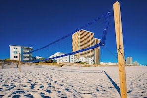A beach volleyball net with a wooden post, sandy beach, and buildings in the background. - Phoenix Gulf Towers Unit 2606 (Orange Beach)