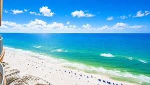 A beach with clear blue water, a sandy shore, and a building with a balcony overlooking the beach. - Phoenix Gulf Towers 2706 (Orange Beach)