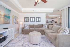 A living room with a sectional sofa, a round ottoman, a TV, and a ceiling fan.