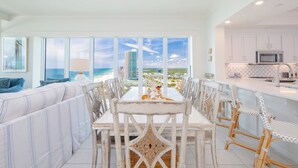 A dining area with a view of the beach and ocean, white furniture, and a kitchen with white cabinets.
