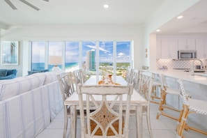 A dining area with a view of the beach and ocean, white furniture, and a kitchen with white cabinets.