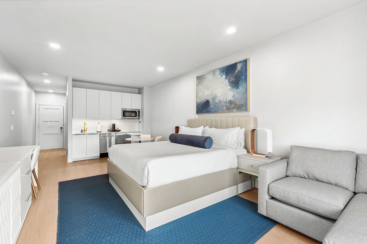 A modern hotel room with a large bed, a grey sofa, a desk with a lamp, and a kitchen area in the background.
