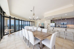 Un vaste espace de salle à manger avec une grande table en bois et des chaises blanches, une cuisine équipée d’appareils électroménagers modernes et un balcon offrant une vue sur l’océan.