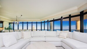 Amplia sala de estar con un sofá seccional blanco grande, una mesa redonda de centro y vista al horizonte de la ciudad a través de amplias ventanas.