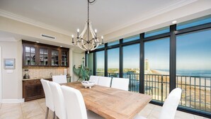 Un comedor con mesa de madera y sillas blancas, una araña y vistas al océano a través de grandes ventanales.