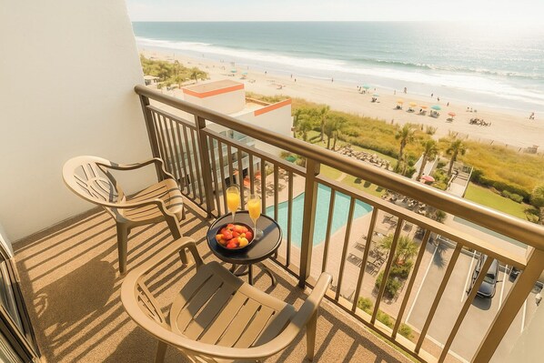 A balcony with a view of the beach, a pool, and a table with fruit and drinks.