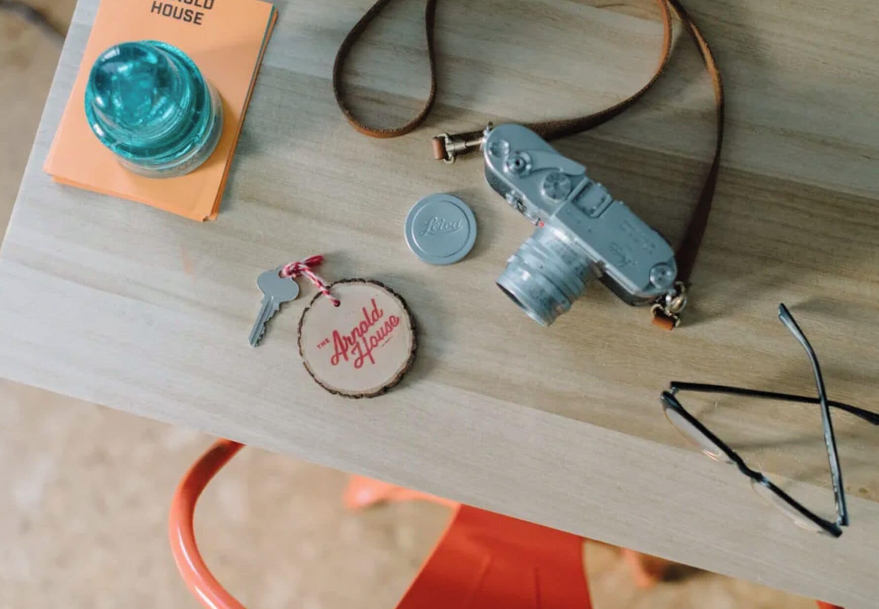 A table with a vintage camera, a keychain, a pair of glasses, a blue bottle, and a small orange book titled "THE ARNOLD HOUSE".