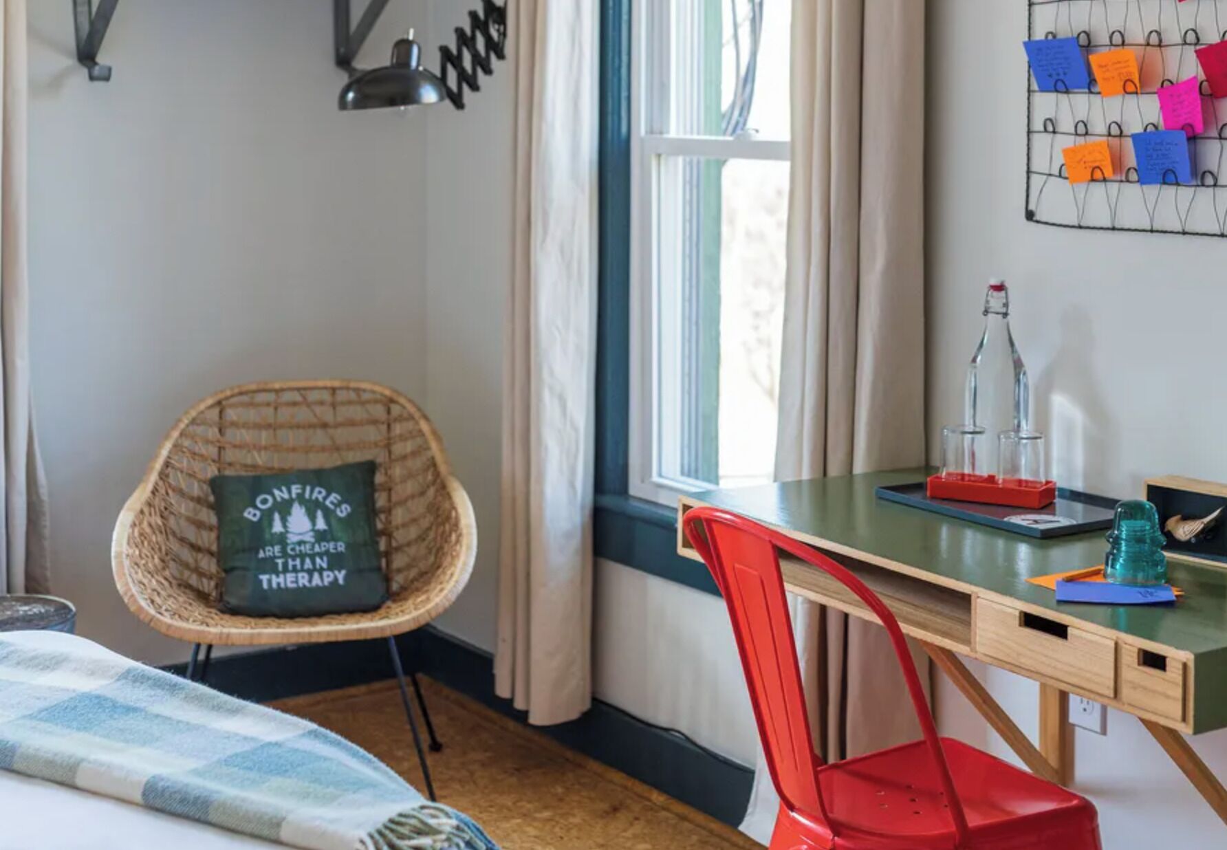 A room with a wicker chair, a red chair, a wooden desk, and a window with curtains.
