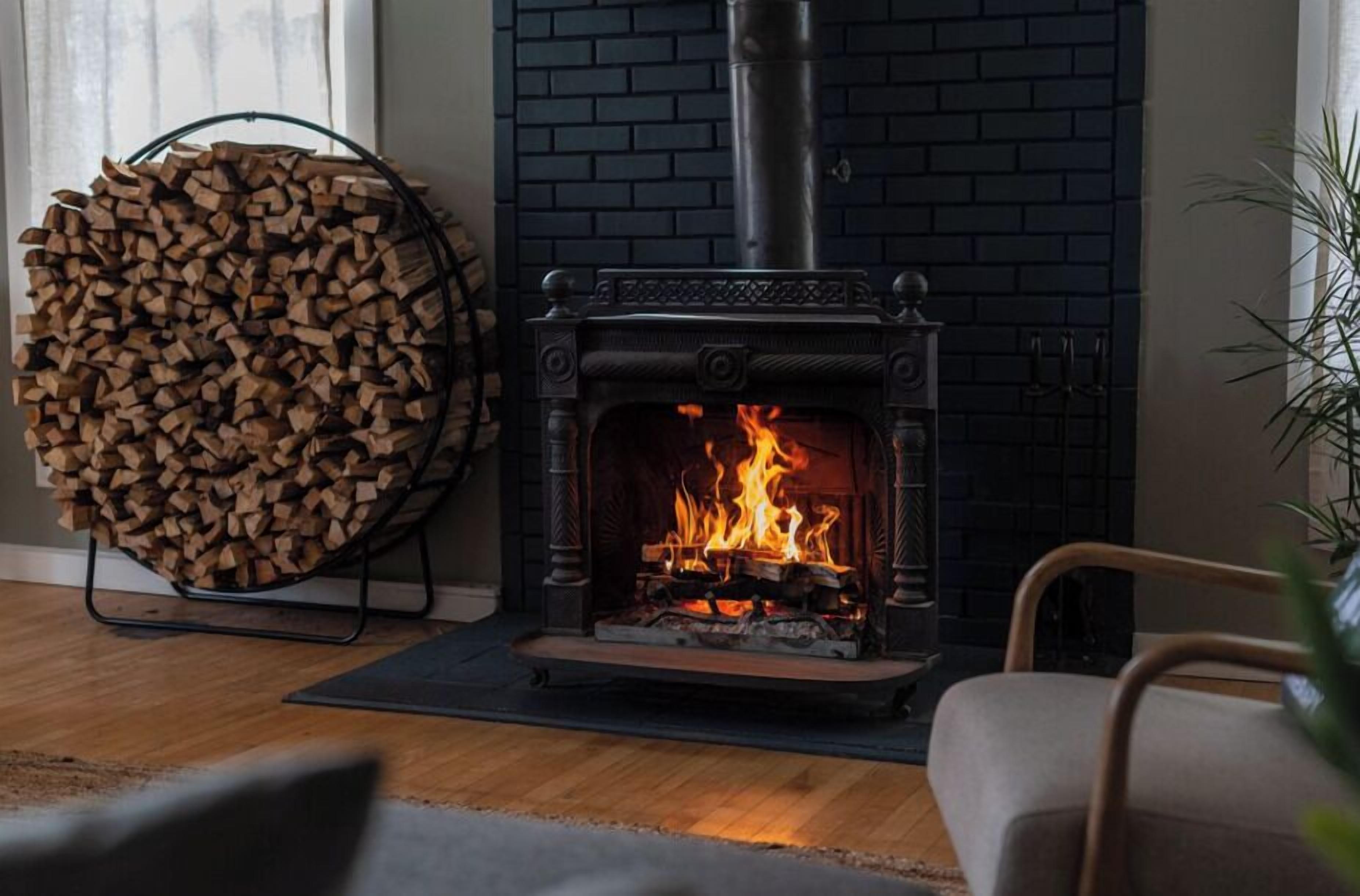 En vedeldad kamin med en brasa som sprakar inuti, en hög med ved och en stol i ett rum med en tegelvägg.