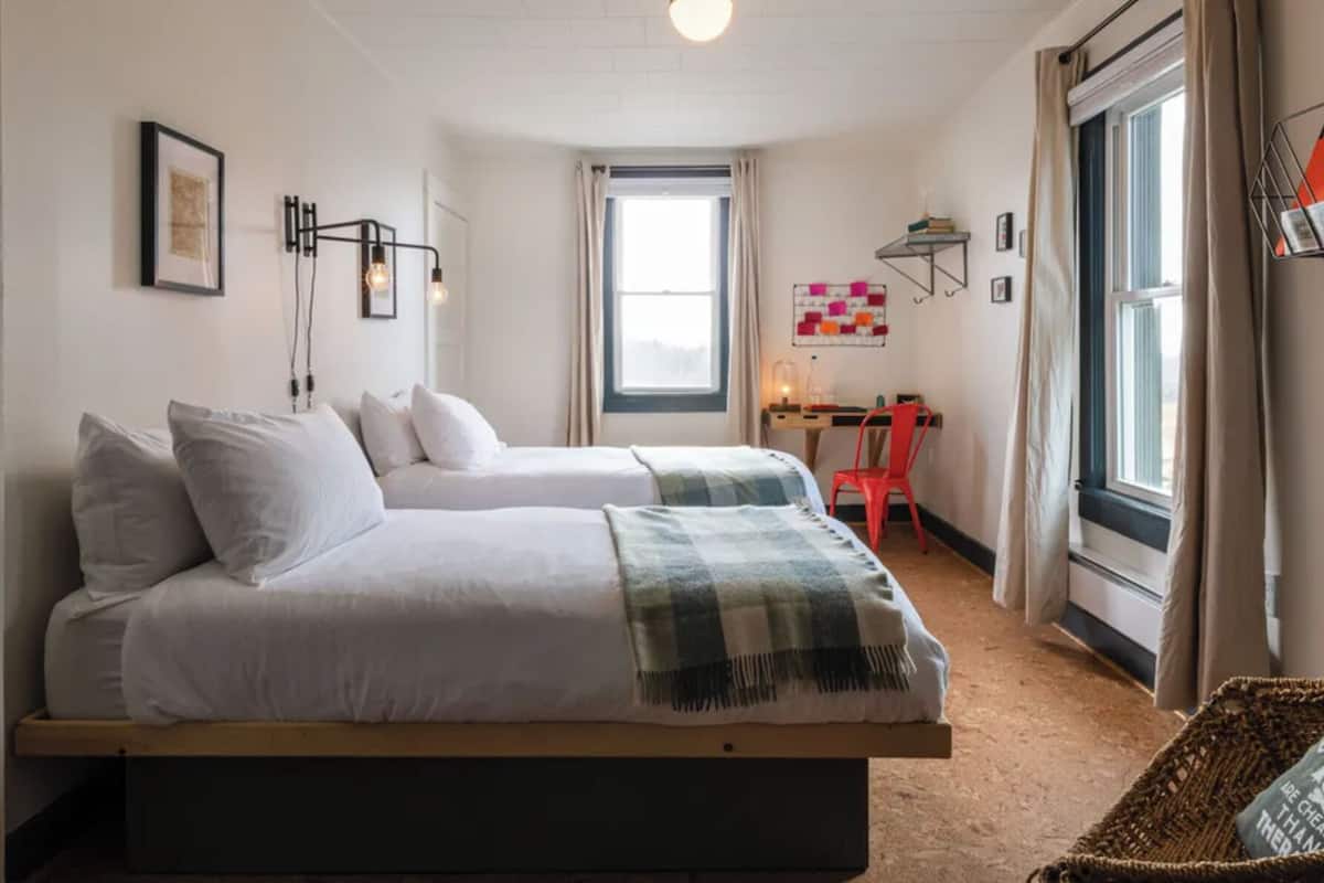 A bedroom with two beds, a desk, and a chair. There is a window with curtains, a wall-mounted light fixture, and a framed picture.