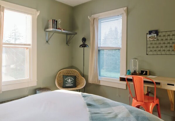 Um quarto com cama, escrivaninha, cadeira, janela e estante com livros.