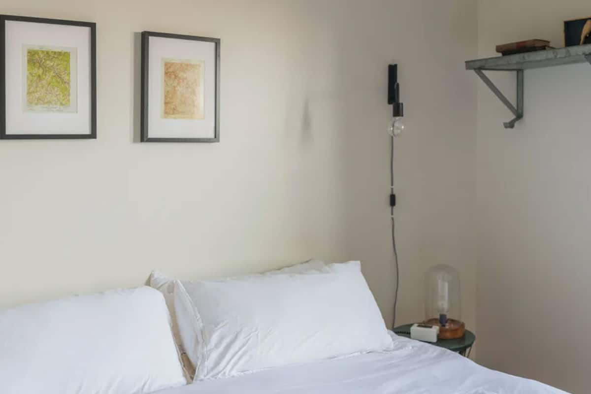 A bed with white linens, a bedside table with a lamp, and two framed artworks on the wall.