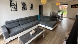 A modern living room with a grey sectional sofa, a coffee table, and a dining area with a table and chairs. - Villa-Iska-Calme-Piscine-Moderne-Port-Centre-Plages (La Tremblade)