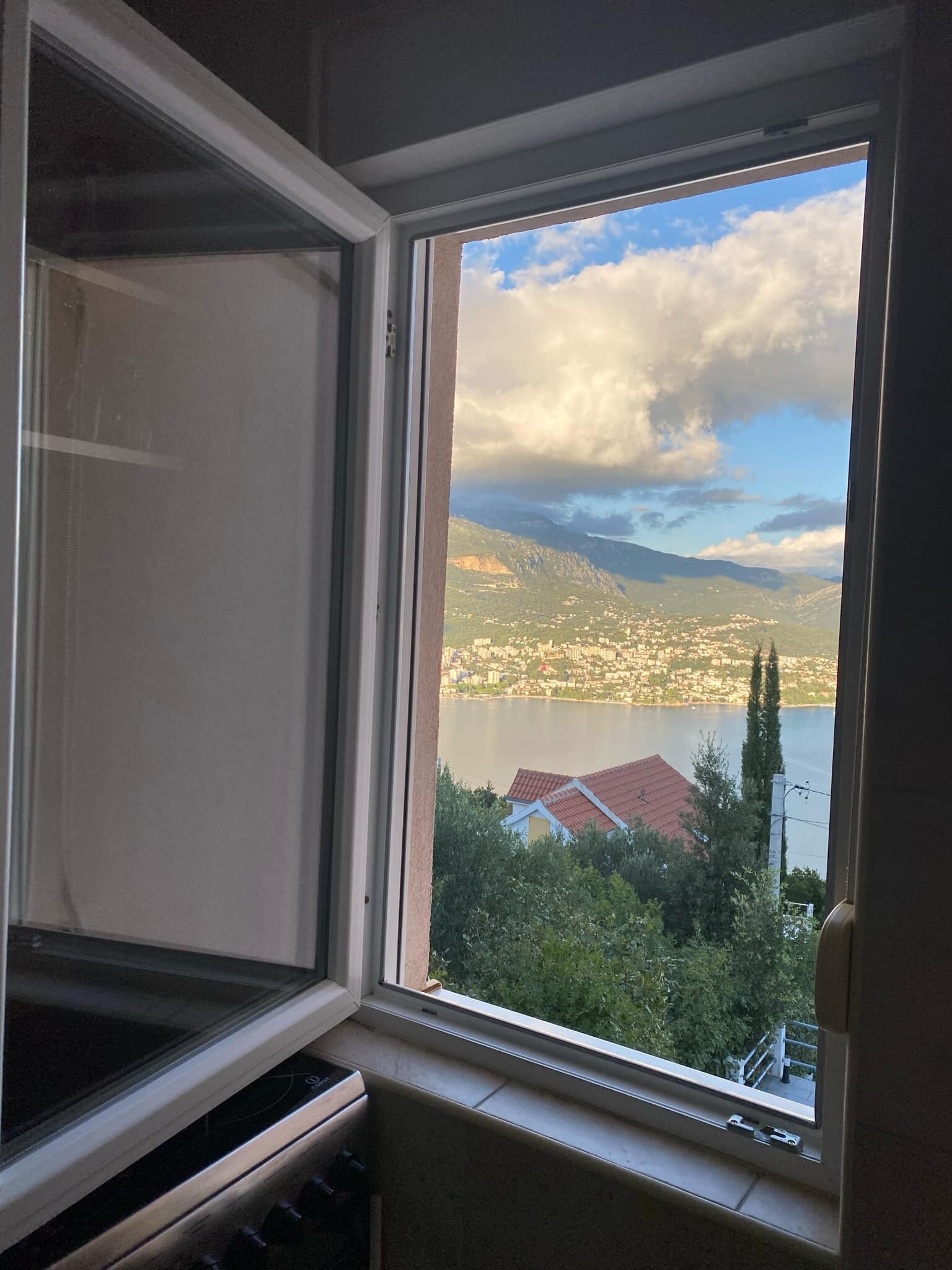 Une fenêtre donnant sur un paysage urbain, des montagnes et une étendue d’eau.