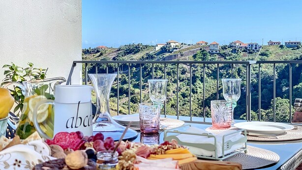 A balcony with a table set for a meal, overlooking a hillside with houses. - Sevenhouse by Atlantic Holiday (Machico)