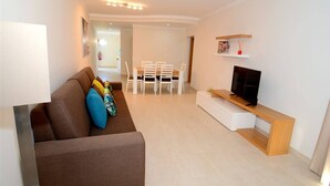 Un salon moderne avec un canapé marron, un téléviseur posé sur un meuble en bois et un coin repas comprenant des chaises blanches et une table.