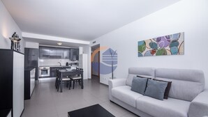 A modern living room with a grey sofa, a coffee table, and a dining area with a table and chairs. There is a kitchen in the background with cabinets and appliances. - 1 Bedroom apartment Paraiso Sol da Rocha (Portimão)