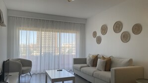 A modern living room with a sofa, a coffee table, and a TV. There are curtains on the window, and wall decor is visible. - 1 Bedroom apartment Paraiso Sol da Rocha (Portimão)