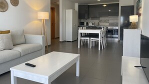 A modern living room with a white sofa, a white coffee table, and a dining area with a white table and chairs. A kitchen is visible in the background with dark cabinets and a microwave. - 1 Bedroom apartment Paraiso Sol da Rocha (Portimão)