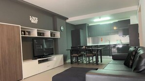 En moderne stue med et fladskærms-tv, en sofa, et spisebord med stole, og et køkken i baggrunden.