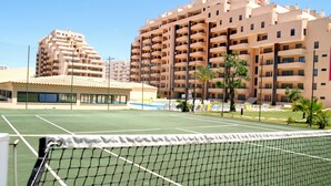 Ein Tennisplatz mit Netz im Vordergrund, umgeben von einem großen, mehrstöckigen Gebäude mit Balkonen.