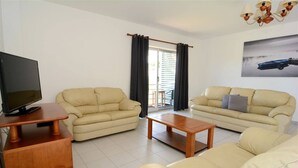 Un salon avec deux canapés beige, une table basse en bois, un téléviseur posé sur un meuble et un balcon avec vue.