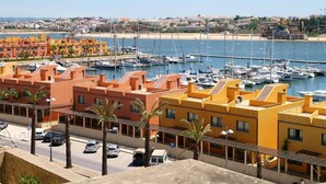 Un port de plaisance situé sur la côte, avec des bâtiments aux couleurs vives, un ciel dégagé et une plage de sable en arrière-plan.