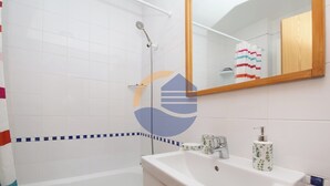 Ein Badezimmer mit weißen Fliesen, einer Dusche, einem Waschbecken mit Armatur, einem Spiegel und einem Handtuchhalter.