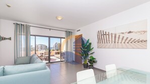 A modern living room with a glass table, a sofa, and a large window with curtains. - 3 Bedroom apartment Paraiso Sol da Rocha (Portimão)