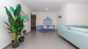 A modern living room with a light blue sofa, a dining area with a table and chairs, and two large potted plants. - 3 Bedroom apartment Paraiso Sol da Rocha (Portimão)