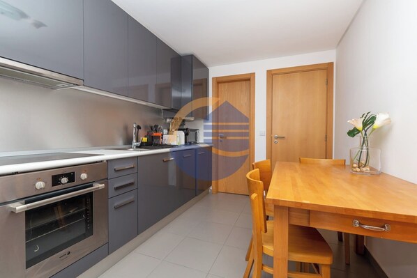 A modern kitchen with grey cabinets, a built-in oven, and a wooden dining table with chairs. - 3 Bedroom apartment Paraiso Sol da Rocha (Portimão)