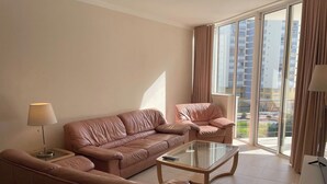 A modern living room with a brown leather sofa, a glass coffee table, and a view of high-rise buildings through large windows. - 2 Bedroom apartment Condominio Primavera (Portimão)