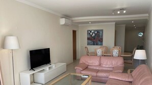 A modern living room with a flat-screen TV, a glass coffee table, a pink sofa, and a dining area with chairs and a table. - 2 Bedroom apartment Condominio Primavera (Portimão)