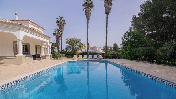 Ein Swimmingpool mit Fliesenrand, ein Gebäude mit Säulen und Palmen.