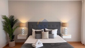Une chambre à coucher moderne avec une tête de lit capitonnée, deux tables de chevet avec des lampes, une grande plante en pot et des serviettes soigneusement pliées sur le lit.