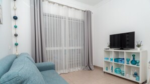 A living room with a sofa, a TV on a shelf, and a window with sheer curtains.