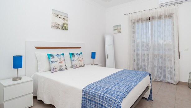 A bedroom with a bed, nightstands, a lamp, a blue blanket, and a window with curtains.