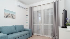 A modern living room with a blue sofa, a large window with sheer curtains, a flat-screen TV, and a wall-mounted air conditioner.