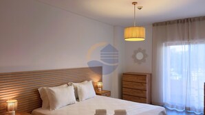 A hotel room with a bed, bedside lamps, a wooden headboard, a nightstand, a wardrobe, and a large window with sheer curtains. - 1 Bedroom apartment Paraiso Sol da Rocha (Portimão)