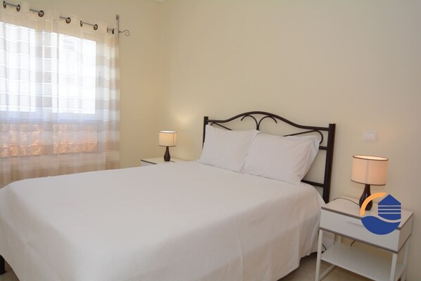 Una cama bien hecha con sábanas blancas, dos lámparas de noche y una ventana con cortinas translúcidas.
