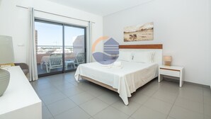 A modern hotel room with a large bed, a bedside table with a lamp, a view of the city through a sliding glass door, and a painting on the wall. - One bedroom Duplex Paraíso Sol da Rocha (Portimão)