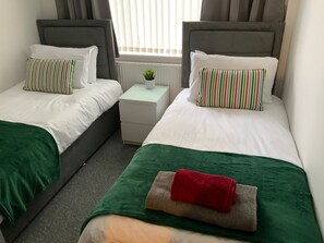 A twin room with two beds, each with a green blanket and striped pillows. There is a small bedside table with a plant and a white lamp.