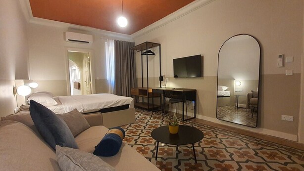 A hotel room with a bed, a sofa, a desk, a chair, a mirror, and a small table with a plant. - Capital Central Suites No2 - Peppa (Valletta, Malta,)