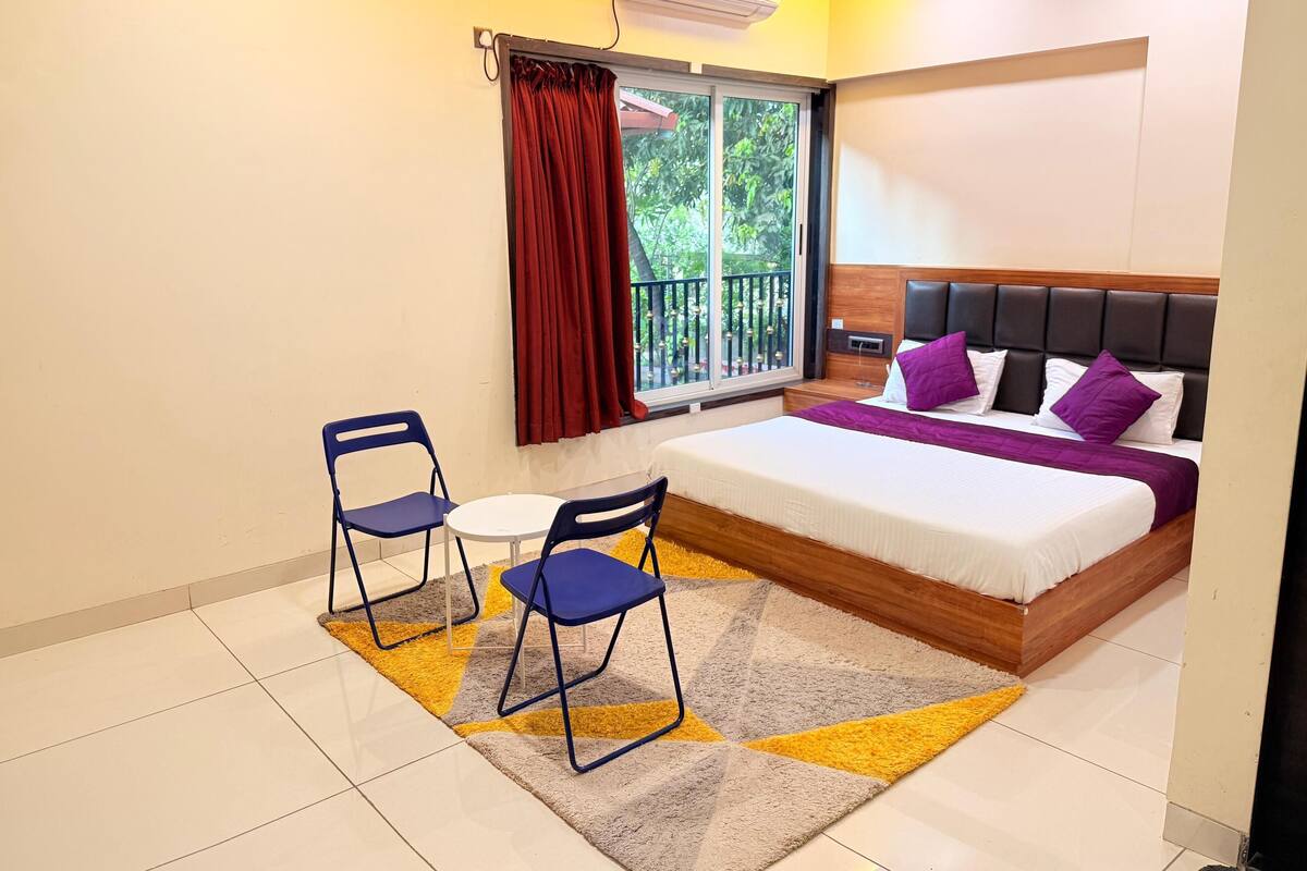 A bedroom with a bed, two chairs, a small table, a window with curtains, and an air conditioning unit.