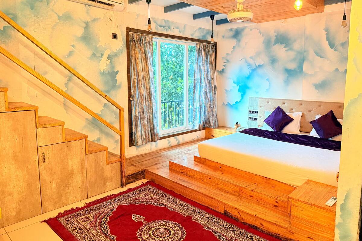 A bedroom with a bed, a window with curtains, a wooden staircase, and a red patterned rug.