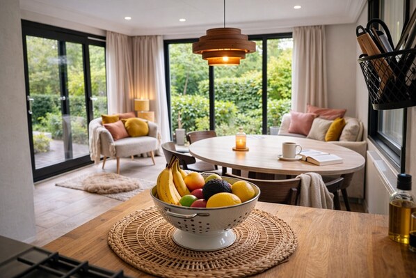 A dining area with a table, a bowl of fruit, and a view of greenery through large windows.